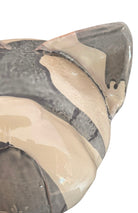 A ceramic sculpture of a bulldog's head by Enza Fasano. The piece features a glossy, glazed finish with a sophisticated color palette of muted greys, creams, and earthy brown tones arranged in bold, abstract striped patterns across the face and ears. The bulldog has characteristic folded ears, a prominent snout, and a soulful, slightly downturned expression. The sculpture is set against a clean, white background, highlighting its smooth texture and reflective surface.