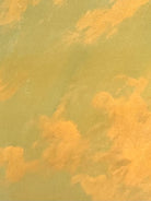 An oil painting on a wooden panel depicts a solitary scarecrow standing in a vast, grassy field under a pale yellow and blue sky. The scarecrow is shown from a three-quarters back view, wearing a brimmed hat and a tattered, patched brown coat with frayed straw bursting from its sleeves and edges. In the far left background, a long, white rustic barn sits before a dense line of green trees. The entire piece is enclosed in a thick, weathered, natural wood frame.