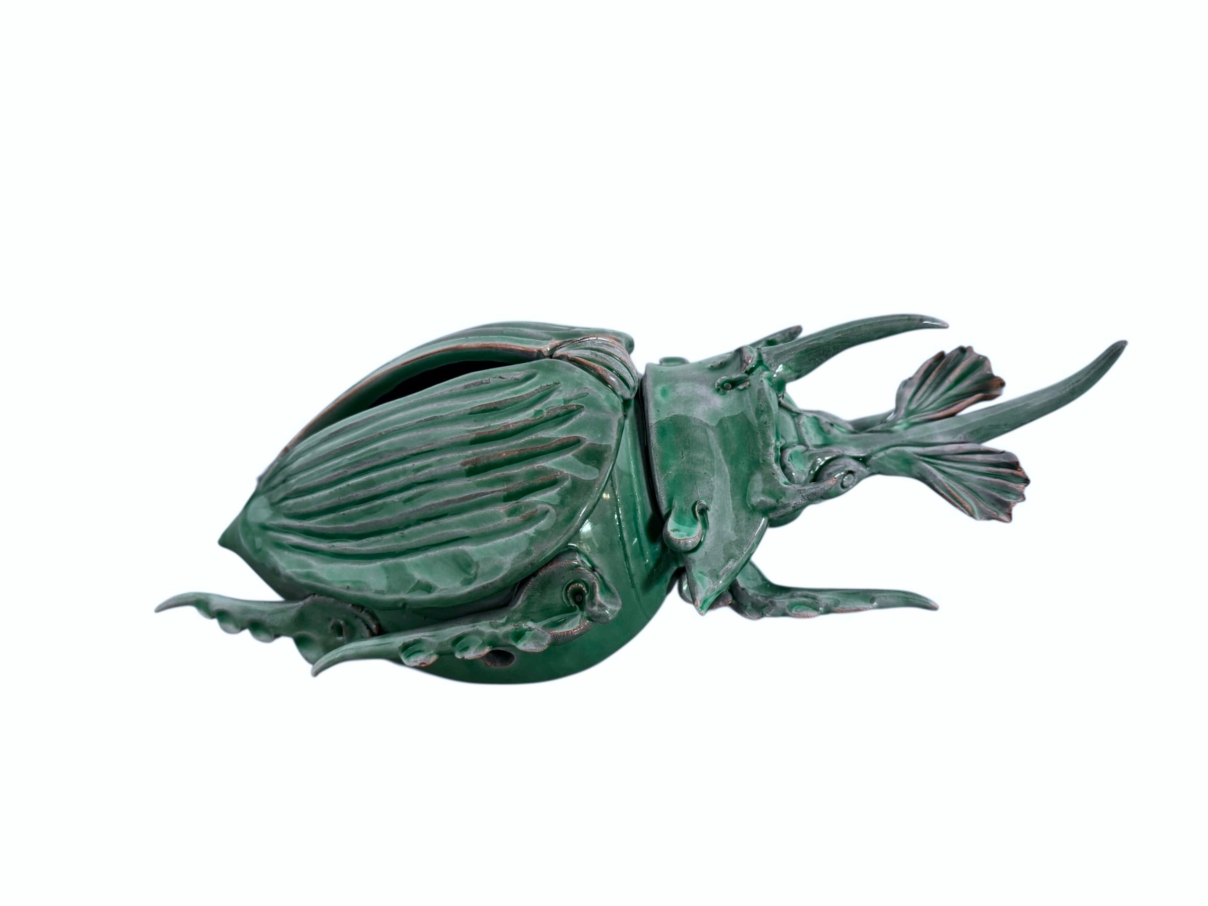 A ceramic sculpture of a stylized beetle by Enza Fasano, featuring a deep emerald green glaze with dark, metallic bronze-colored accents. The beetle is depicted with a highly textured, ribbed shell (elytra) and prominent, curved horns. Its legs are stylized with suckers, reminiscent of octopus tentacles. The piece has a glossy finish and rests against a plain white background.