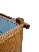 A three-tiered wooden shelving unit featuring a mix of light oak and teal-colored panels. The piece has an open, structural design with visible joinery, vertical supports that extend past the top shelf, and a horizontal backrest at the top. The middle and bottom shelves feature slatted wood center sections flanked by solid teal panels, while the top surface is primarily solid wood with teal accents at the edges.