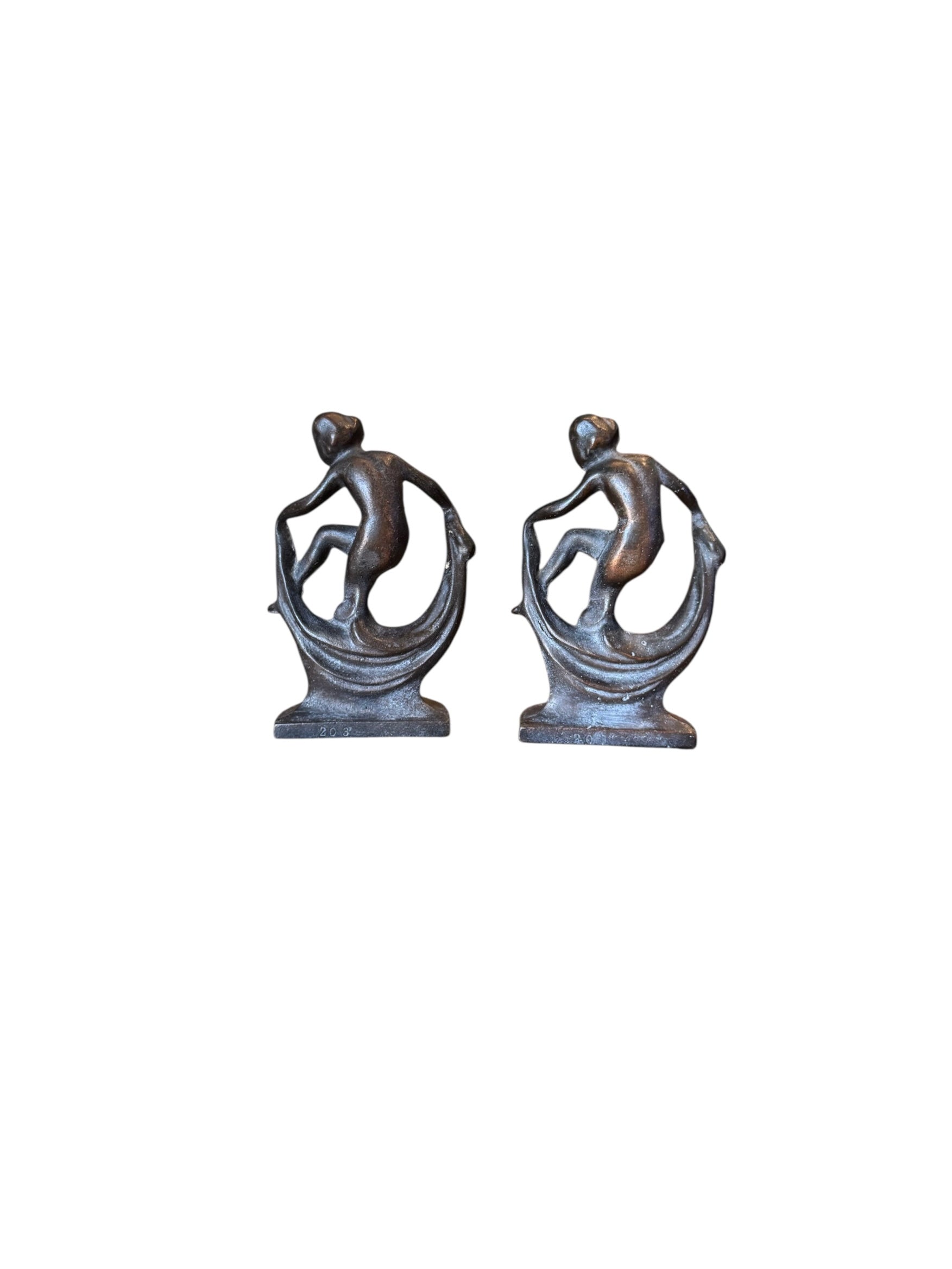 Two cast iron bookends in the Art Deco style. Each features a nude female figure leaning gracefully with one leg extended, posed atop a stylized wave or drape. The bookends have a bronze-colored finish and a vintage appearance.