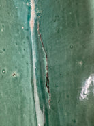 curved ceramic piece resembling a green pod, supported by a block plinth. The seafoam-green ceramic structure extends wide on either side, curving upward to form a wavy, boat-like shape. The outer surface is dotted with rows of small, raised, brown-orange, acorn-like protrusions. The top rim is layered with parallel, grooved ridges. The base is a vertical rectangular block with a heavily textured, bark-like surface in a dark green and black finish.