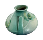 A squat, rounded vintage Roseville pottery vase in a light teal-blue matte glaze. The vase features two small, angular geometric handles on its shoulders and a flared rim. Embossed on the front is a cluster of textured, green and pale-yellow thornapple blossoms tucked against sharp, serrated green leaves. The surface shows a subtle, fine network of age-related crazing lines throughout the glaze.