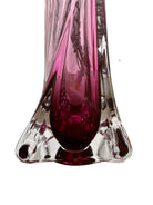 A tall, Mid-Century Modern sculptural table lamp featuring a hand-blown art glass base. The base is crafted in the Sommerso style, with a deep amethyst purple core encased in a thick, clear crystal outer layer that twists upward in an organic, fluid motion. The lamp is topped with a white, knife-pleated empire shade and a clear glass ball finial. The overall design is elegant and slender, emphasizing the contrast between the saturated purple glass and the transparent, light-refracting crystal.