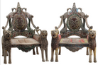 A pair of antique, heavily carved Anglo-Indian style throne chairs featuring lion figures as legs and arm supports, and peacock motifs on the backrests. The chairs have a distressed, multi-colored painted finish.