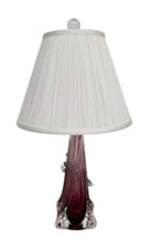 A tall, Mid-Century Modern sculptural table lamp featuring a hand-blown art glass base. The base is crafted in the Sommerso style, with a deep amethyst purple core encased in a thick, clear crystal outer layer that twists upward in an organic, fluid motion. The lamp is topped with a white, knife-pleated empire shade and a clear glass ball finial. The overall design is elegant and slender, emphasizing the contrast between the saturated purple glass and the transparent, light-refracting crystal.