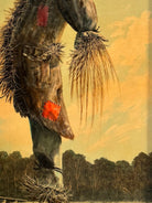 An oil painting on a wooden panel depicts a solitary scarecrow standing in a vast, grassy field under a pale yellow and blue sky. The scarecrow is shown from a three-quarters back view, wearing a brimmed hat and a tattered, patched brown coat with frayed straw bursting from its sleeves and edges. In the far left background, a long, white rustic barn sits before a dense line of green trees. The entire piece is enclosed in a thick, weathered, natural wood frame.