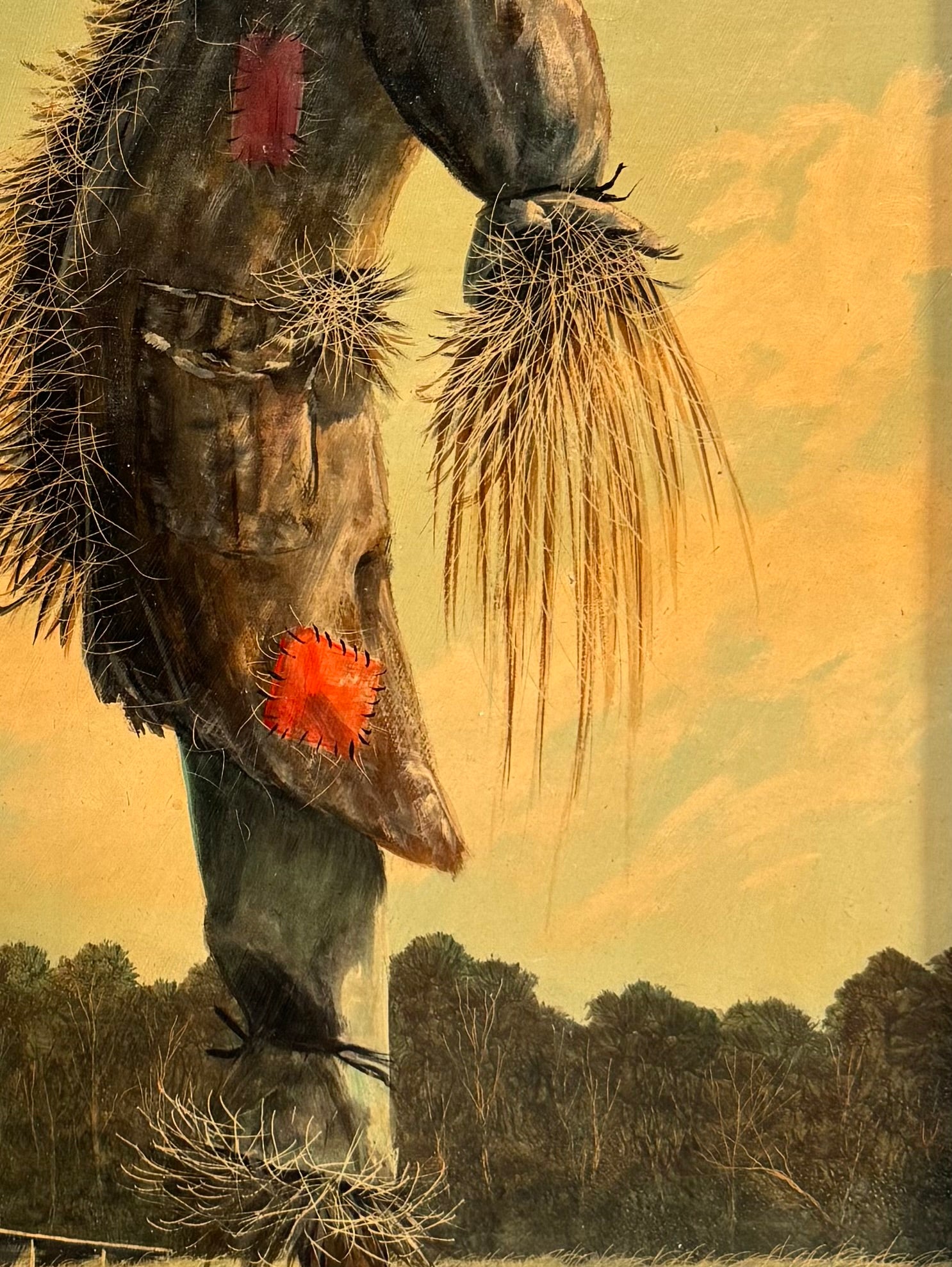 An oil painting on a wooden panel depicts a solitary scarecrow standing in a vast, grassy field under a pale yellow and blue sky. The scarecrow is shown from a three-quarters back view, wearing a brimmed hat and a tattered, patched brown coat with frayed straw bursting from its sleeves and edges. In the far left background, a long, white rustic barn sits before a dense line of green trees. The entire piece is enclosed in a thick, weathered, natural wood frame.