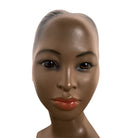 a vintage ceramic bust featuring a woman with brown skin, dark eyes, and orange lips. Her black hair is sleek and parted on the side, pulled back into a low style. She wears a small, round white earring in her right ear lobe. The bust includes her head, neck, and the upper part of her shoulders