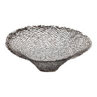 A round metal wall sculpture by Michael Aram, viewed from the front against a stark white background. The piece is composed of a dense, intricate lattice of fine, dark metal wire or twigs woven together to create a textured, web-like pattern. The structure curves inward like a funnel or a vortex, sloping down toward a clean, circular opening in the exact center, creating a strong sense of depth and dimensionality.