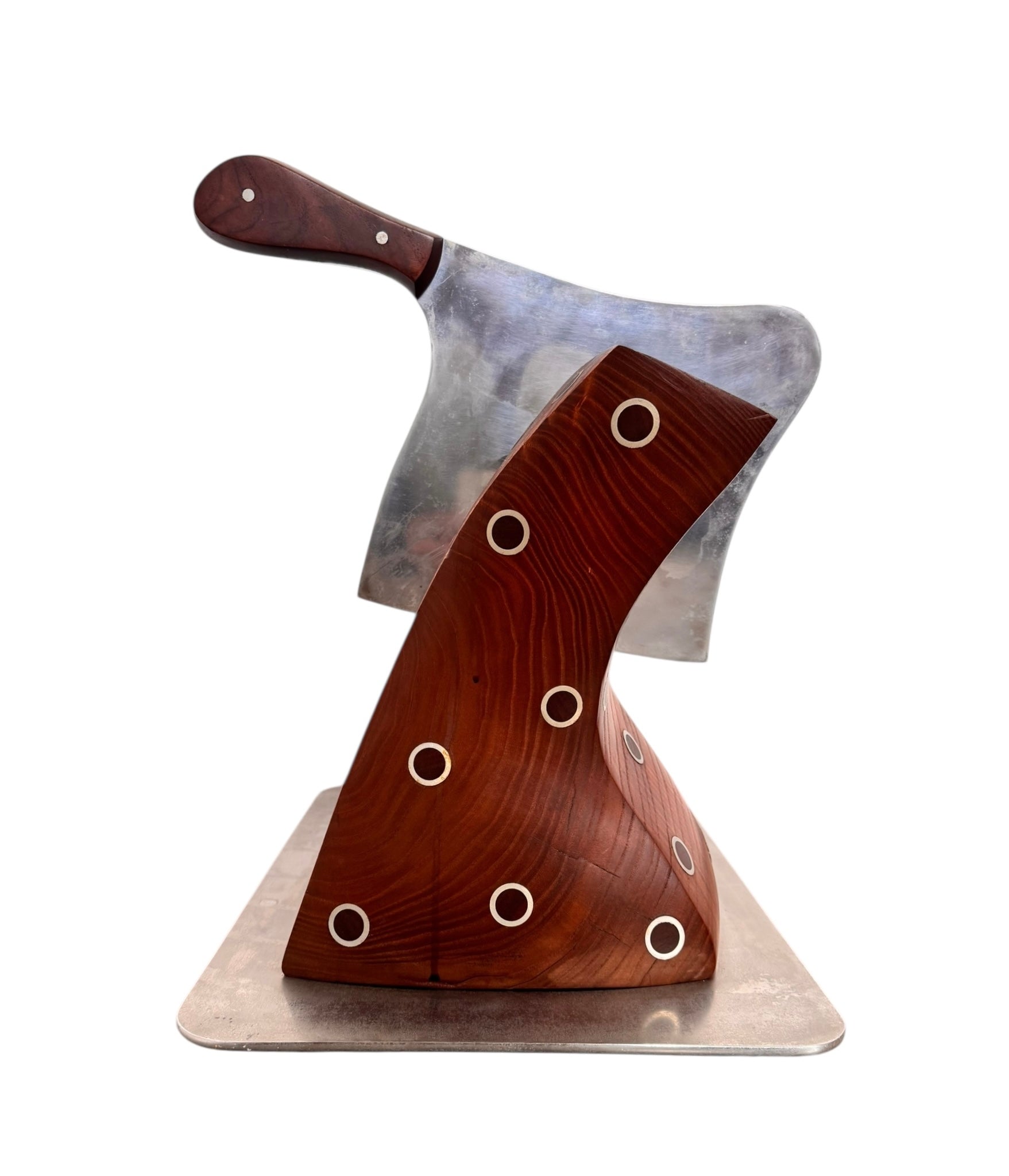 A wooden sculpture with a highly polished, reddish-brown grain, featuring two main asymmetrical, curved sections that converge at the top. Circular metal inlays with black centers are embedded across the surface of both wooden sections. A flat, polished metal blade-like element is inserted vertically between the two wooden sections, extending above the top surface. The sculpture rests on a metallic base.