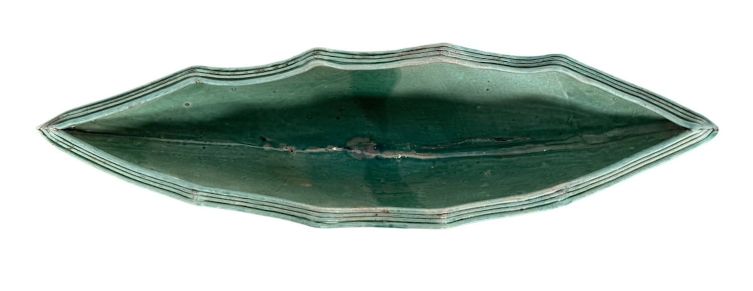 curved ceramic piece resembling a green pod, supported by a block plinth. The seafoam-green ceramic structure extends wide on either side, curving upward to form a wavy, boat-like shape. The outer surface is dotted with rows of small, raised, brown-orange, acorn-like protrusions. The top rim is layered with parallel, grooved ridges. The base is a vertical rectangular block with a heavily textured, bark-like surface in a dark green and black finish.