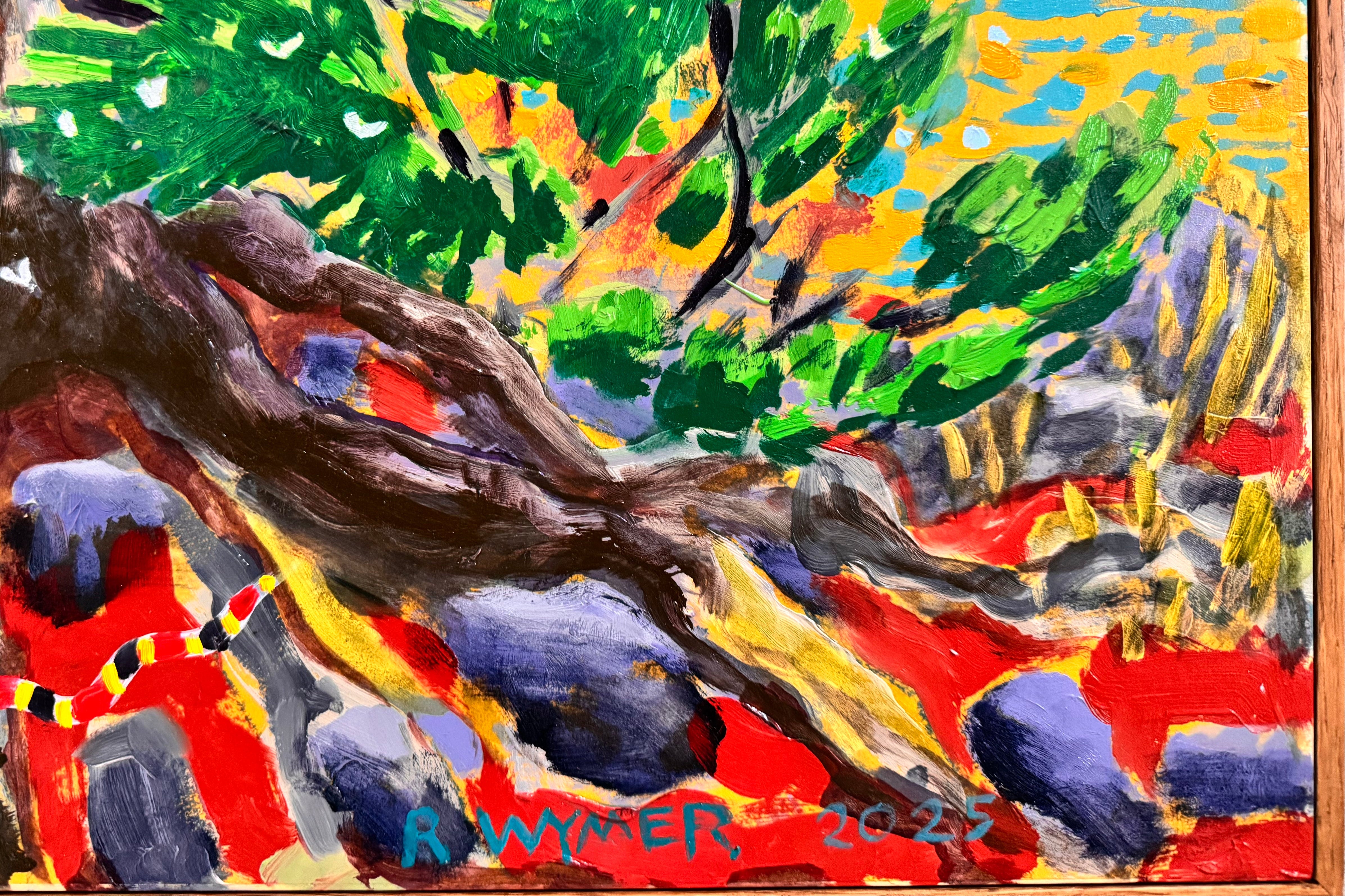 A vibrantly colored landscape painting titled 'Perdernales Falls Fisherman,' featuring two large trees with prominent exposed roots framing a body of water. A lone figure, a fisherman, stands by the water in the middle ground. The painting uses bold strokes of yellow, green, red, and blue, and is enclosed in a rustic dark wooden frame with a light-colored inner mat.