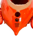 A glossy, vibrant orange ceramic sculpture of a rhinoceros beetle by Enza Fasano. The piece features a highly polished, reflective glaze and stylized, fluid anatomical details. Its prominent dual horns curve forward, and the body is textured with layered, wing-like plates, set against a plain white background.