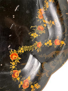 A vintage rectangular toleware serving tray with a scalloped, wavy edge and a black lacquered finish. The tray features hand-painted floral motifs in a traditional folk-art style. At the bottom center is a large, vibrant cluster of yellow and orange roses and daisies with green leaves. Smaller floral sprays and delicate gold filigree scrollwork decorate the corners and the upper rim. The center of the tray is open black space, showing light surface wear and minor scuffing consistent with age.