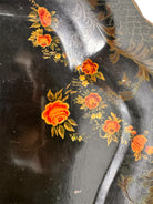 A vintage rectangular toleware serving tray with a scalloped, wavy edge and a black lacquered finish. The tray features hand-painted floral motifs in a traditional folk-art style. At the bottom center is a large, vibrant cluster of yellow and orange roses and daisies with green leaves. Smaller floral sprays and delicate gold filigree scrollwork decorate the corners and the upper rim. The center of the tray is open black space, showing light surface wear and minor scuffing consistent with age.