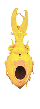 a large, high-gloss ceramic sculpture by Enza Fasano, shaped like a stylized beetle. The entire piece is finished in a warm, honey-yellow glaze. The beetle features prominent, curved mandibles at the front, a textured "collar" area, and a rounded, shell-like body with decorative ridges along the edges. The surface is smooth and reflective, catching the light across its organic contours.