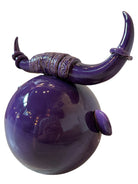 Glossy purple ceramic sculpture featuring a round body and stylized bull horns adorned with rings, and a nose ring. The sculpture has no eyes, but suggests a smiling expression.