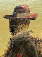 An oil painting on a wooden panel depicts a solitary scarecrow standing in a vast, grassy field under a pale yellow and blue sky. The scarecrow is shown from a three-quarters back view, wearing a brimmed hat and a tattered, patched brown coat with frayed straw bursting from its sleeves and edges. In the far left background, a long, white rustic barn sits before a dense line of green trees. The entire piece is enclosed in a thick, weathered, natural wood frame.