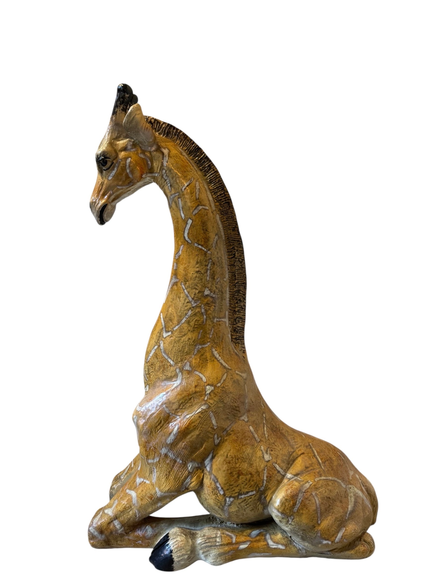 A hand-painted, cast resin sculpture of a seated giraffe. The giraffe has a long neck, is predominantly gold with light markings, and is in a resting pose. The sculpture is against a plain white background