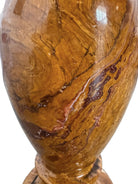 An elegant, hand-carved decorative vase made of polished Pakistani brown onyx. The vase features a classic, rounded urn shape with a tiered, flared neck and a solid matching spherical stone stopper resting on top. The stone displays rich, natural horizontal banding and swirling patterns in shades of honey, deep amber, rust-red, and cream. It stands on a small, integrated pedestal base against a clean white background, reflecting a high-gloss, mirror-like finish.