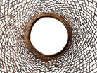 A round metal wall sculpture by Michael Aram, viewed from the front against a stark white background. The piece is composed of a dense, intricate lattice of fine, dark metal wire or twigs woven together to create a textured, web-like pattern. The structure curves inward like a funnel or a vortex, sloping down toward a clean, circular opening in the exact center, creating a strong sense of depth and dimensionality.