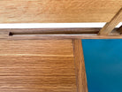A three-tiered wooden shelving unit featuring a mix of light oak and teal-colored panels. The piece has an open, structural design with visible joinery, vertical supports that extend past the top shelf, and a horizontal backrest at the top. The middle and bottom shelves feature slatted wood center sections flanked by solid teal panels, while the top surface is primarily solid wood with teal accents at the edges.