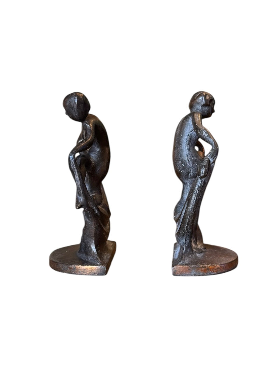 Two cast iron bookends in the Art Deco style. Each features a nude female figure leaning gracefully with one leg extended, posed atop a stylized wave or drape. The bookends have a bronze-colored finish and a vintage appearance.