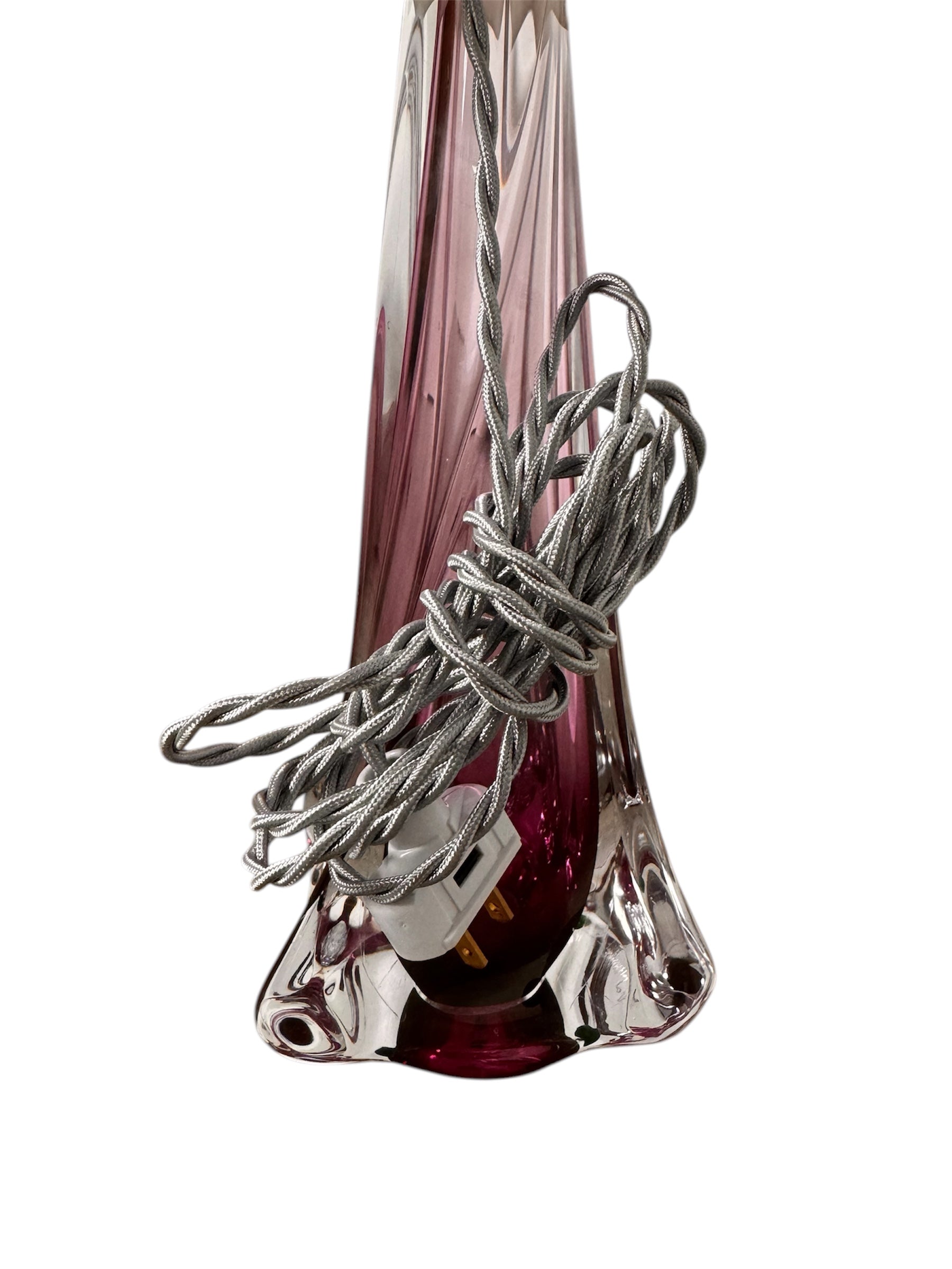 A tall, Mid-Century Modern sculptural table lamp featuring a hand-blown art glass base. The base is crafted in the Sommerso style, with a deep amethyst purple core encased in a thick, clear crystal outer layer that twists upward in an organic, fluid motion. The lamp is topped with a white, knife-pleated empire shade and a clear glass ball finial. The overall design is elegant and slender, emphasizing the contrast between the saturated purple glass and the transparent, light-refracting crystal.