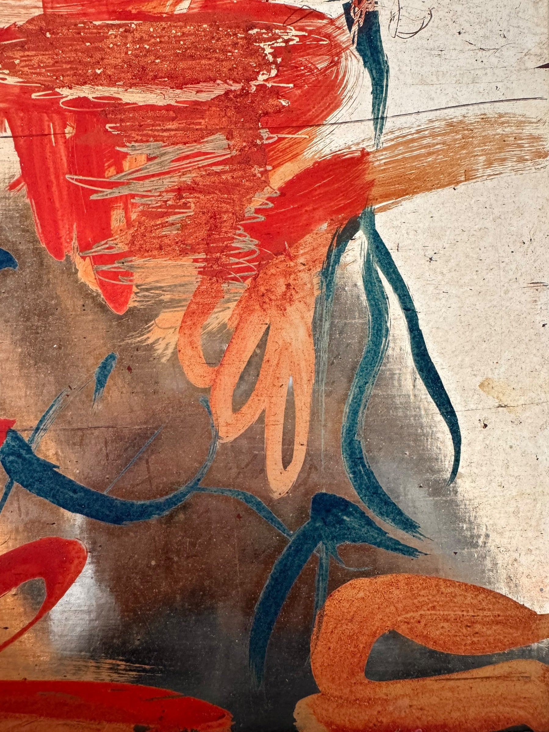 A vertical composition on a bright, reflective metallic background (likely silver or light gold leaf). The piece is defined by sparse, high-energy gestures: a sweeping arc of orange and red at the top, a central cluster of textured red paint, and a scattering of teal-blue "C" shapes and hooks. The metallic surface is highly visible, giving the piece a luminous, modern feel.