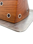 A wooden sculpture with a highly polished, reddish-brown grain, featuring two main asymmetrical, curved sections that converge at the top. Circular metal inlays with black centers are embedded across the surface of both wooden sections. A flat, polished metal blade-like element is inserted vertically between the two wooden sections, extending above the top surface. The sculpture rests on a metallic base.