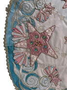 A round, cream-colored tablecloth featuring a dramatic, scalloped border shaped like six large flower petals. The border is intricately embroidered with a coastal motif including pink starfish, seashells, and teal-blue swirling waves. Gold beaded accents line the inner circular seam and the outer edges.
A long, narrow cream runner with matching scalloped, wavy edges. It is adorned with the same nautical embroidery of pink starfish and seashells scattered across the center and concentrated along the edge.
