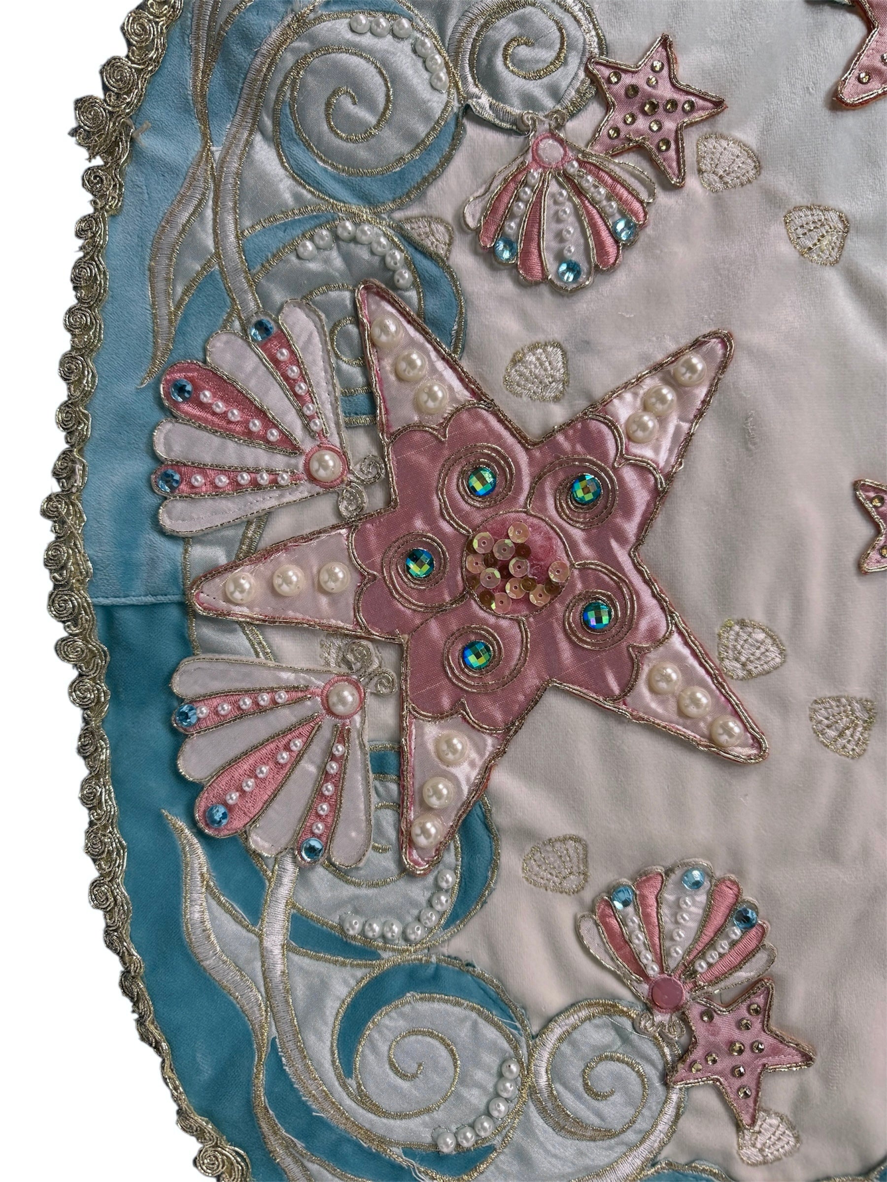 A round, cream-colored tablecloth featuring a dramatic, scalloped border shaped like six large flower petals. The border is intricately embroidered with a coastal motif including pink starfish, seashells, and teal-blue swirling waves. Gold beaded accents line the inner circular seam and the outer edges.
A long, narrow cream runner with matching scalloped, wavy edges. It is adorned with the same nautical embroidery of pink starfish and seashells scattered across the center and concentrated along the edge.