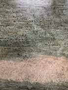 A realistic painting of a rustic interior scene by Travis Whitfield. A duck sits inside a woven basket on a dark wooden table with a single drawer. A French horn hangs on the light blue and white log wall behind the table. A multi-pane window on the left side of the painting shows an outdoor view. 