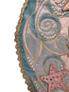 A round, cream-colored tablecloth featuring a dramatic, scalloped border shaped like six large flower petals. The border is intricately embroidered with a coastal motif including pink starfish, seashells, and teal-blue swirling waves. Gold beaded accents line the inner circular seam and the outer edges.
A long, narrow cream runner with matching scalloped, wavy edges. It is adorned with the same nautical embroidery of pink starfish and seashells scattered across the center and concentrated along the edge.