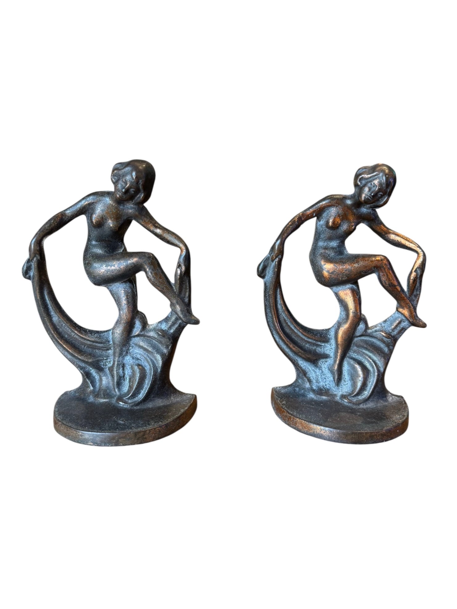 Two cast iron bookends in the Art Deco style. Each features a nude female figure leaning gracefully with one leg extended, posed atop a stylized wave or drape. The bookends have a bronze-colored finish and a vintage appearance.
Bronze Echoes: Art Deco Bookends attributed to Hubley Mold