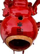 A handcrafted ceramic sculpture of a rhinoceros beetle by Enza Fasano, featuring a vibrant, glossy deep-red glaze. The piece is stylized with smooth, rounded contours and prominent, elegant horns extending forward. Its surface reflects light, highlighting the rich texture and artisanal detail of the clay.