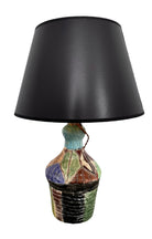 A hand-crafted ceramic table lamp featuring a vibrant, multi-colored base and a sleek black shade. The base is an upcycled artisan vase with a textured, hand-thrown appearance, decorated with geometric panels in shades of mustard yellow, forest green, cobalt blue, and burgundy, separated by thick black outlines. The lamp is topped with a minimalist, black tapered drum shade that provides a sharp contrast to the colorful, eclectic pottery below.
