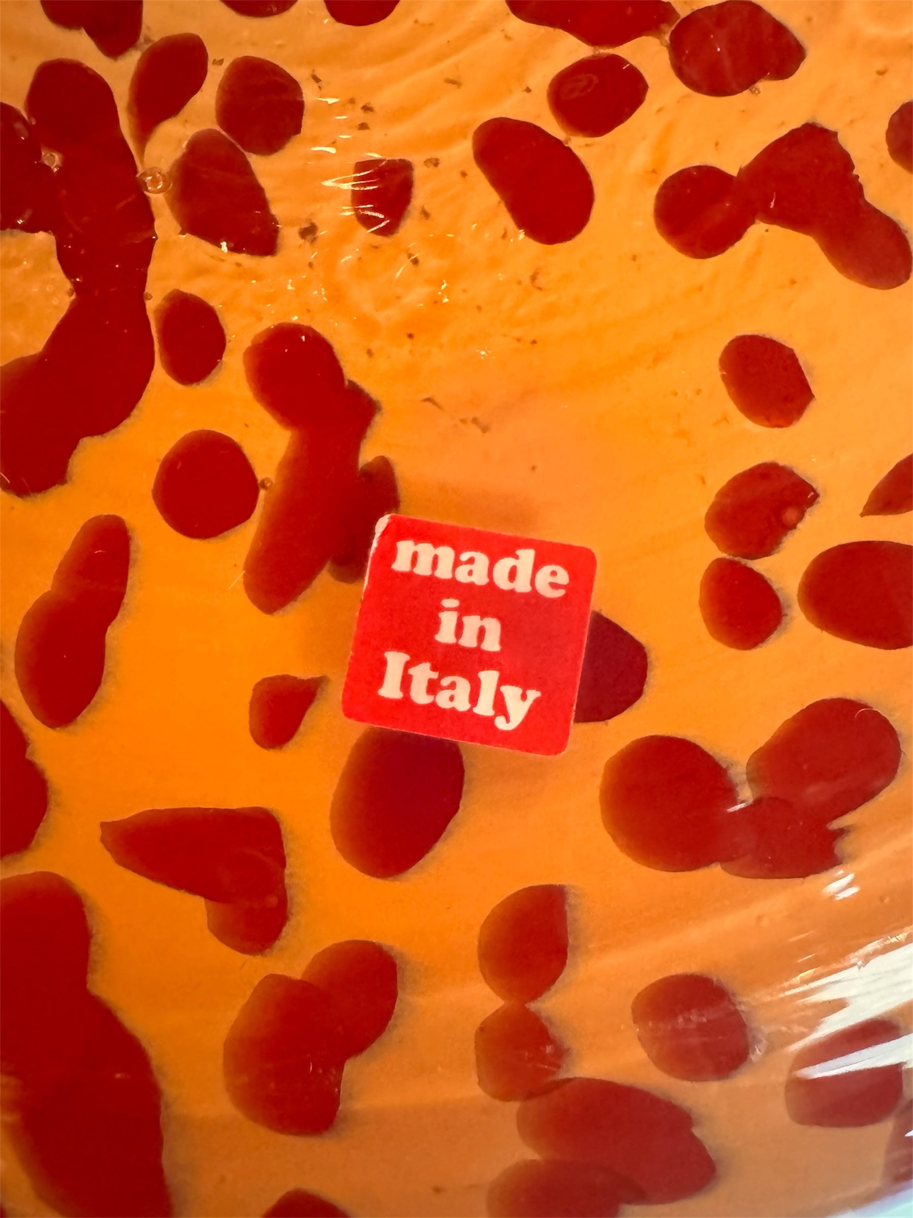 A hand-blown Italian Murano glass vase featuring a wide, bulbous body and a short, flared rim. The glass is a vibrant, translucent amber-orange base decorated with an organic "macchia" or spotted pattern of deep ruby red blotches. The red spots are elongated and vertical near the neck, becoming larger and more rounded toward the base. The surface has a high-gloss, reflective finish, showcasing mid-century modern craftsmanship.
