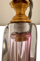 A tall, Mid-Century Modern sculptural table lamp featuring a hand-blown art glass base. The base is crafted in the Sommerso style, with a deep amethyst purple core encased in a thick, clear crystal outer layer that twists upward in an organic, fluid motion. The lamp is topped with a white, knife-pleated empire shade and a clear glass ball finial. The overall design is elegant and slender, emphasizing the contrast between the saturated purple glass and the transparent, light-refracting crystal.