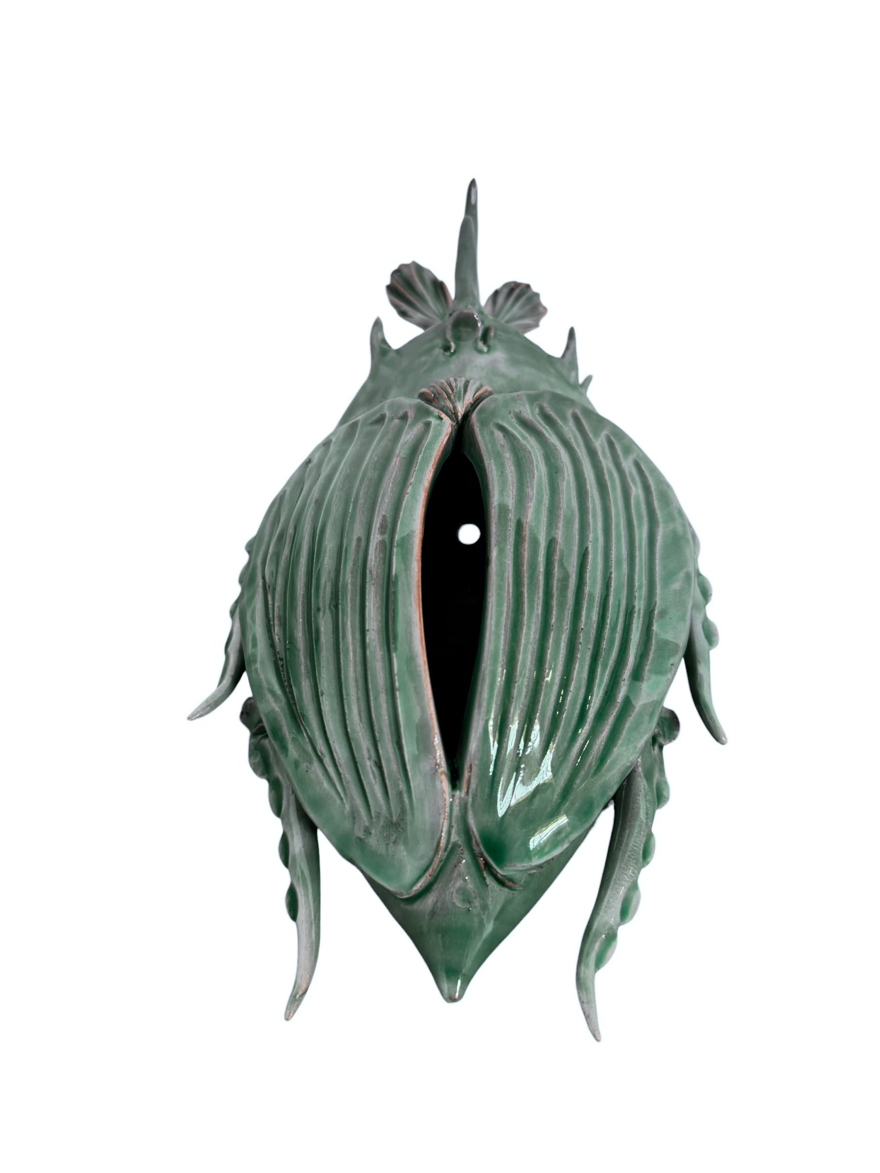 A ceramic sculpture of a stylized beetle by Enza Fasano, featuring a deep emerald green glaze with dark, metallic bronze-colored accents. The beetle is depicted with a highly textured, ribbed shell (elytra) and prominent, curved horns. Its legs are stylized with suckers, reminiscent of octopus tentacles. The piece has a glossy finish and rests against a plain white background.