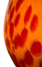 A hand-blown Italian Murano glass vase featuring a wide, bulbous body and a short, flared rim. The glass is a vibrant, translucent amber-orange base decorated with an organic "macchia" or spotted pattern of deep ruby red blotches. The red spots are elongated and vertical near the neck, becoming larger and more rounded toward the base. The surface has a high-gloss, reflective finish, showcasing mid-century modern craftsmanship.
