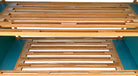 A three-tiered wooden shelving unit featuring a mix of light oak and teal-colored panels. The piece has an open, structural design with visible joinery, vertical supports that extend past the top shelf, and a horizontal backrest at the top. The middle and bottom shelves feature slatted wood center sections flanked by solid teal panels, while the top surface is primarily solid wood with teal accents at the edges.