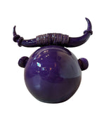 Glossy purple ceramic sculpture featuring a round body and stylized bull horns adorned with rings, and a nose ring. The sculpture has no eyes, but suggests a smiling expression.