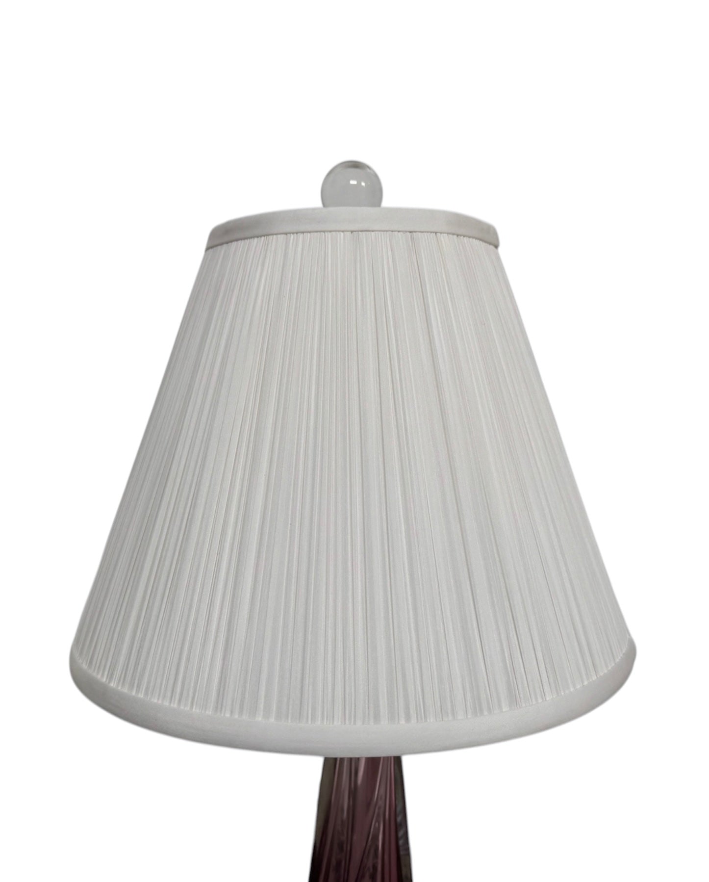A tall, Mid-Century Modern sculptural table lamp featuring a hand-blown art glass base. The base is crafted in the Sommerso style, with a deep amethyst purple core encased in a thick, clear crystal outer layer that twists upward in an organic, fluid motion. The lamp is topped with a white, knife-pleated empire shade and a clear glass ball finial. The overall design is elegant and slender, emphasizing the contrast between the saturated purple glass and the transparent, light-refracting crystal.