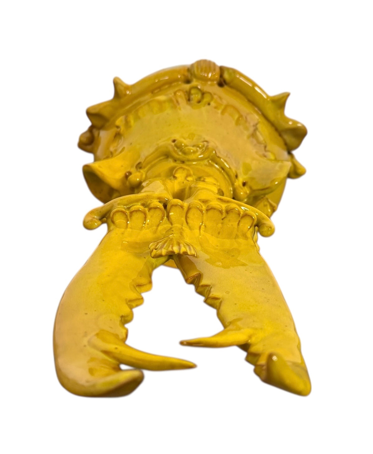 a large, high-gloss ceramic sculpture by Enza Fasano, shaped like a stylized beetle. The entire piece is finished in a warm, honey-yellow glaze. The beetle features prominent, curved mandibles at the front, a textured "collar" area, and a rounded, shell-like body with decorative ridges along the edges. The surface is smooth and reflective, catching the light across its organic contours.