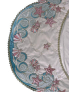 A round, cream-colored tablecloth featuring a dramatic, scalloped border shaped like six large flower petals. The border is intricately embroidered with a coastal motif including pink starfish, seashells, and teal-blue swirling waves. Gold beaded accents line the inner circular seam and the outer edges.
A long, narrow cream runner with matching scalloped, wavy edges. It is adorned with the same nautical embroidery of pink starfish and seashells scattered across the center and concentrated along the edge.