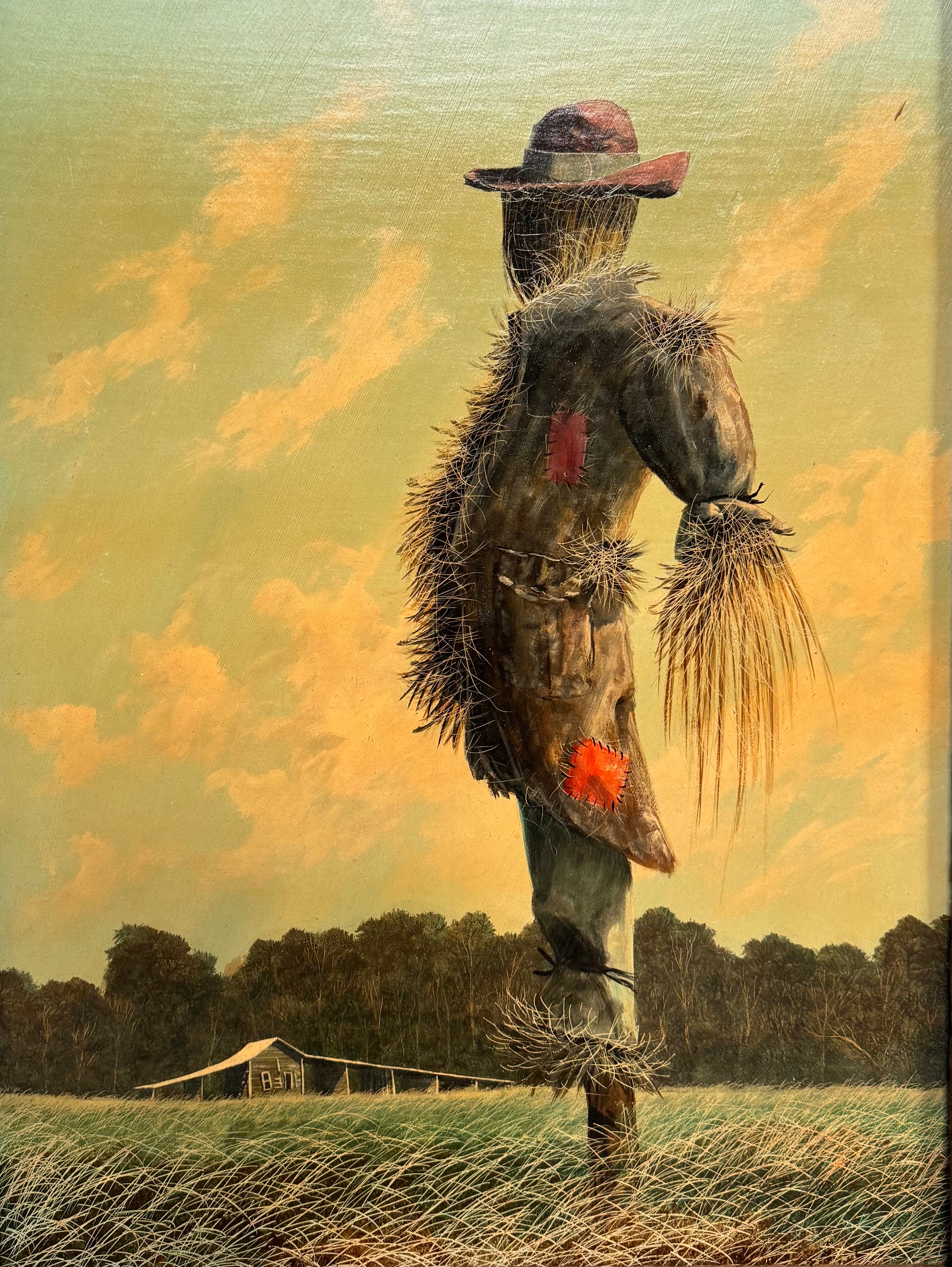 An oil painting on a wooden panel depicts a solitary scarecrow standing in a vast, grassy field under a pale yellow and blue sky. The scarecrow is shown from a three-quarters back view, wearing a brimmed hat and a tattered, patched brown coat with frayed straw bursting from its sleeves and edges. In the far left background, a long, white rustic barn sits before a dense line of green trees. The entire piece is enclosed in a thick, weathered, natural wood frame.