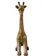 A hand-painted, cast resin sculpture of a seated giraffe. The giraffe has a long neck, is predominantly gold with light markings, and is in a resting pose. The sculpture is against a plain white background