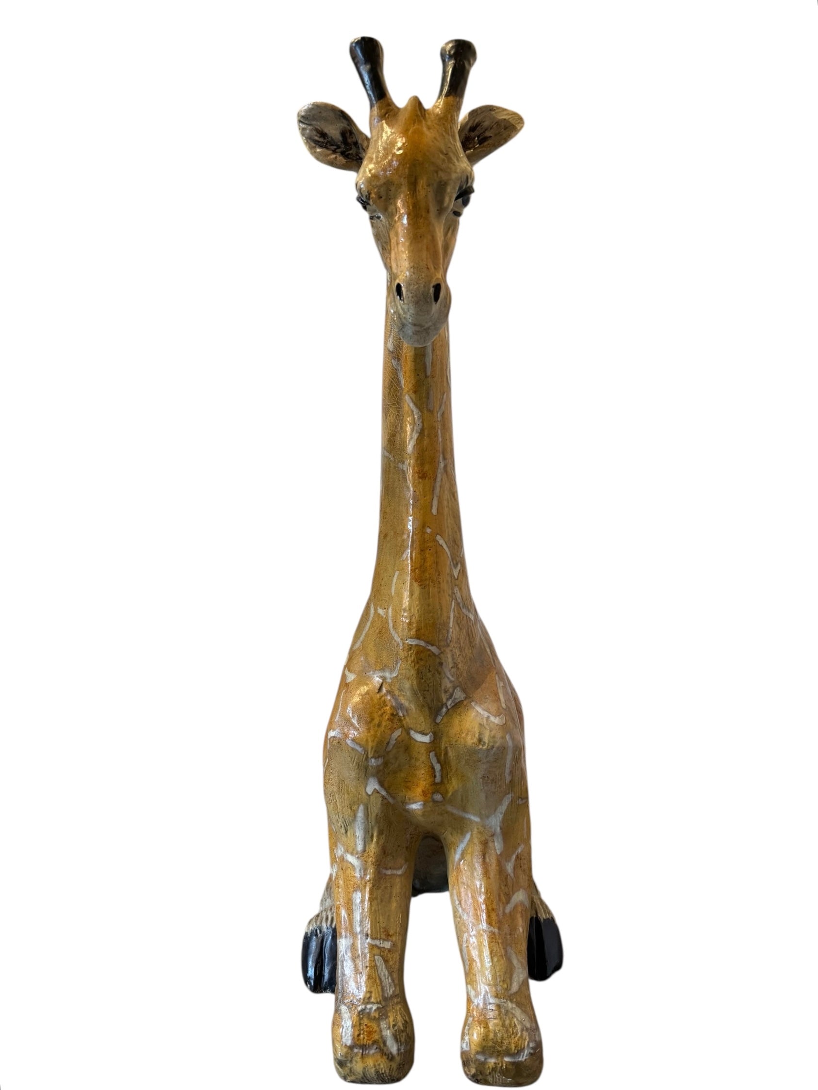 A hand-painted, cast resin sculpture of a seated giraffe. The giraffe has a long neck, is predominantly gold with light markings, and is in a resting pose. The sculpture is against a plain white background